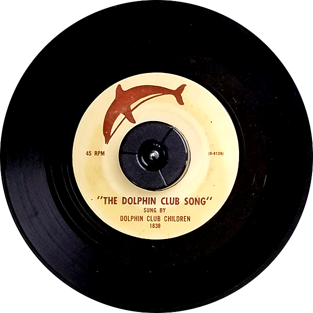 A 45 RPM record with a label in tan and brown color. Featuring a silhouette of a dolphin on the top. The code D-8129 to the right. The bottom has "The Dolphin Club Song sung by Dolphin Club Children" and a number "1838"