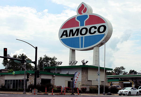 The image shows a large Amoco gas station sign towering above a gas station building. The sign is oval-shaped with a prominent red, white, and blue logo featuring a stylized torch flame above the word "AMOCO" written in bold black letters. The sign is elevated on tall, white pillars, making it a dominant feature in the landscape. Below the sign is a modern gas station with a green and white color scheme, typical of BP-branded stations, indicating that this might be a station that carries both Amoco and BP branding. The surrounding area includes a traffic light, a few vehicles, and some roadwork cones, suggesting a typical urban or suburban setting. The sky is partly cloudy, providing a neutral backdrop to the scene.