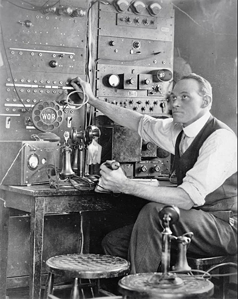 Black and white image depicts a man seated at a control panel filled with various dials, switches, and cables, likely in a radio station or early broadcast facility. He is wearing a vest over a collared shirt with rolled-up sleeves, indicating he is actively engaged in his work. The man is holding a cable or connector with one hand while adjusting a switch or dial with the other. A vintage-style telephone sits on the desk in front of him, along with other broadcasting equipment. A sign with the letters "WOR" is visible on one of the devices, suggesting this might be a historic image related to WOR, a well-known radio station. The scene reflects the early days of radio broadcasting technology.