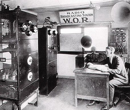 Black and white image shows a man seated at a desk in an early radio broadcasting station. The room is filled with large pieces of equipment, including a tall rack of electronic components and dials on the left, and a console with switches and meters near the man's desk. Above the desk, a large sign reads "Radio Broadcasting Station W.O.R.," clearly identifying the station. The man is dressed in a suit and is either working with the equipment or taking notes. The setting reflects the technological environment of an early 20th-century radio station, highlighting the complexity and scale of the equipment used during that era.