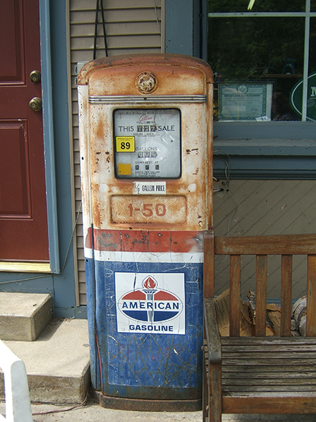 The image shows an old, rusted gas pump located outside a building. The pump is weathered, with visible rust and fading paint, indicating its age. It has a rectangular display with the words "THIS SALE" visible in the top section, and the number "89" in a yellow square, possibly indicating the octane rating of the gasoline. Below the display, there is a large label that reads "AMERICAN GASOLINE" with a logo featuring a flame or torch above the text. The pump is painted in red, white, and blue, though the colors are faded and worn. The gas pump is situated next to a wooden bench and in front of a building with a window and a red door. The scene evokes a sense of nostalgia, reflecting a piece of mid-20th-century Americana.