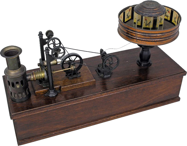 The image shows a vintage mechanical device known as a praxinoscope, an early form of animation technology. The device is mounted on a wooden base and consists of several intricate metal gears and pulleys connected to a cylindrical drum on the right side. The drum has slits around its sides and contains a strip of sequential images inside. When the drum is spun, and the viewer looks through the slits, the images appear to animate, creating the illusion of motion. The left side of the device features a lamp or light source that would illuminate the images, enhancing the visual effect. The overall design is intricate and showcases the craftsmanship of early animation devices.