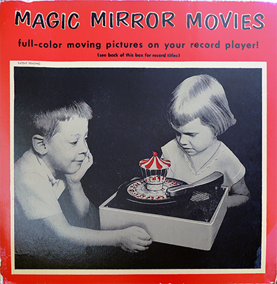 The image shows the cover of a vintage record box for "Magic Mirror Movies." The cover features a black-and-white photograph of two children, a boy and a girl, eagerly watching a Red Raven Magic Mirror in action on a record player. The Magic Mirror, with its colorful circus-like top, is spinning on the record player, bringing the images on the record to life. The title "Magic Mirror Movies" is prominently displayed at the top in bold, red letters, with the subtitle "full-color moving pictures on your record player!" The cover also includes a note to "see back of this box for record titles." The overall design evokes a sense of nostalgia, highlighting the charm and appeal of this early form of home entertainment.