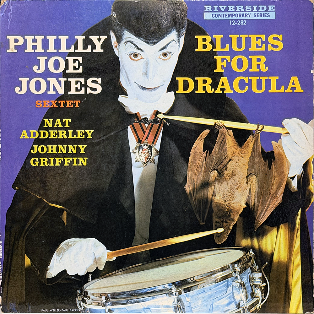 Album cover of Blues for Dracula (1958) by the Philly Joe Jones Sextet, released on Riverside Records. The artwork shows Philly Joe Jones dressed as Dracula with pale makeup, slicked black hair, a cape, and a medallion. He holds a pair of drumsticks, one of which suspends a bat over a snare drum. The background is purple with bold yellow and white text listing the album title, Jones’ name, and the featured musicians Nat Adderley and Johnny Griffin.